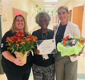 3 women holding flowers and a certificate honoring sub of the month who is standing in the middle